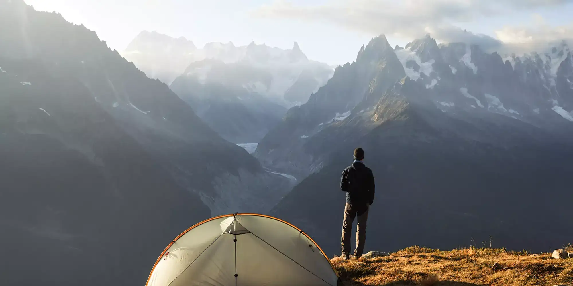 A beige dome tent with a tan trim stands on a grassy mountain ledge.