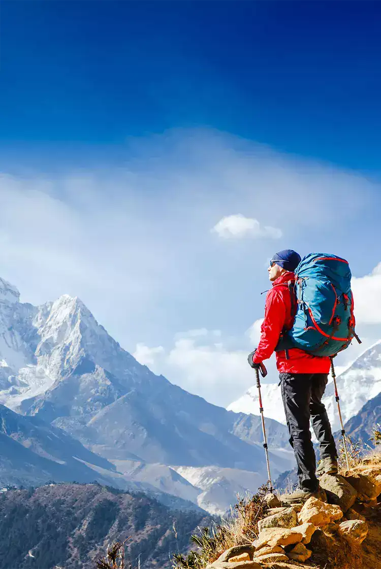 Blue hiking backpack with red straps worn by a person in a red jacket standing on rocky terrain.