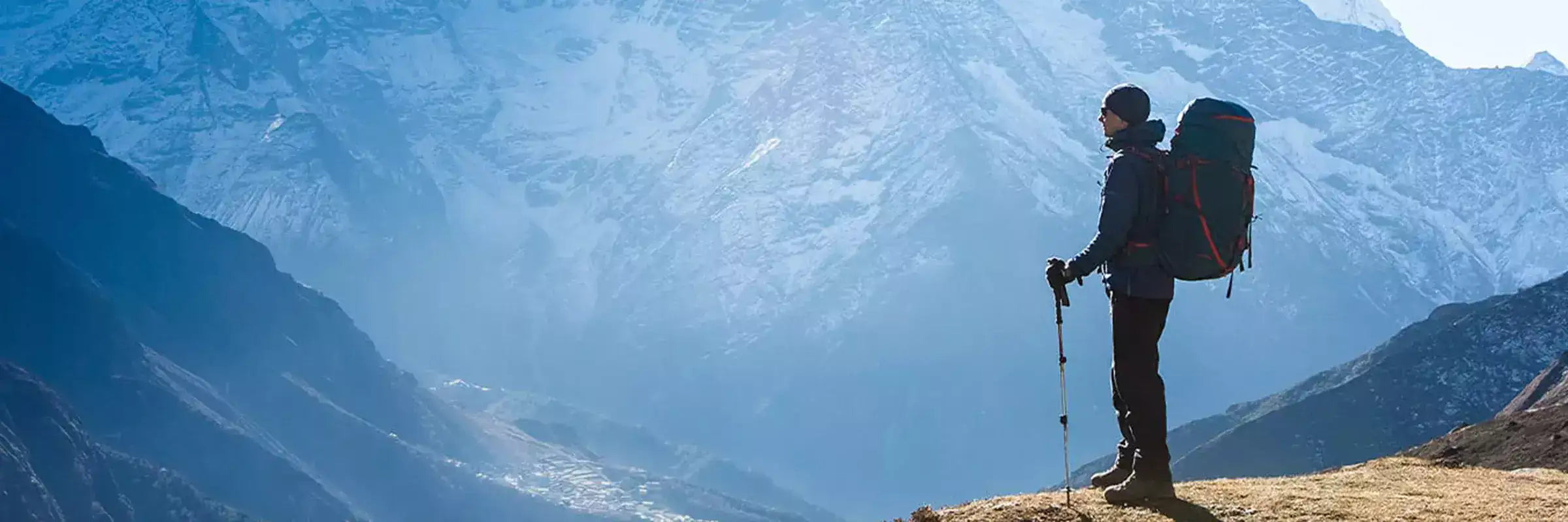 A hiker stands on a mountain ridge wearing a dark jacket and pants, carrying a large black backpack with red trim.